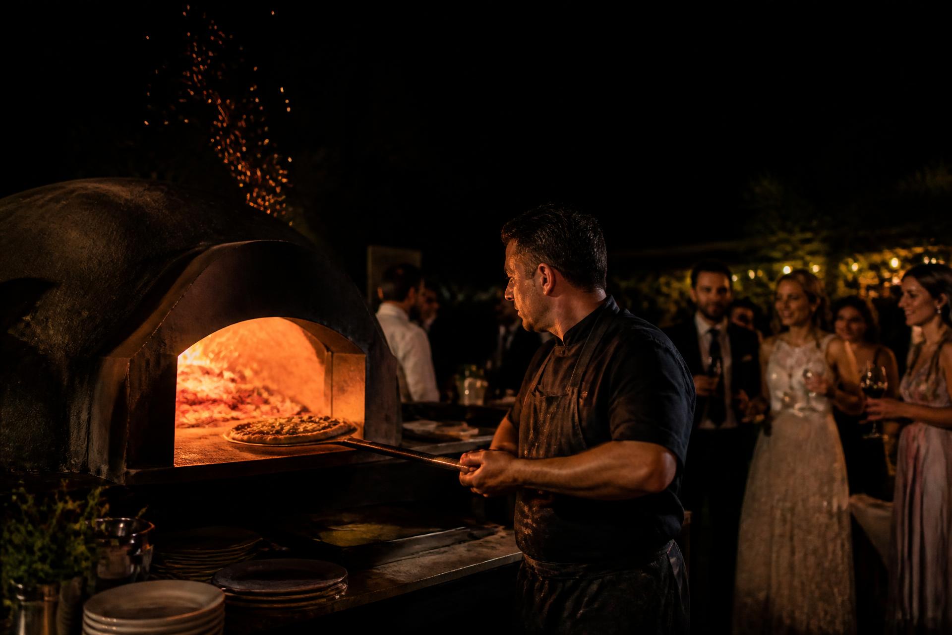 Pizzaiolo sacando una pizza del horno de leña en mitad de una boda de noche, con los invitados alrededor