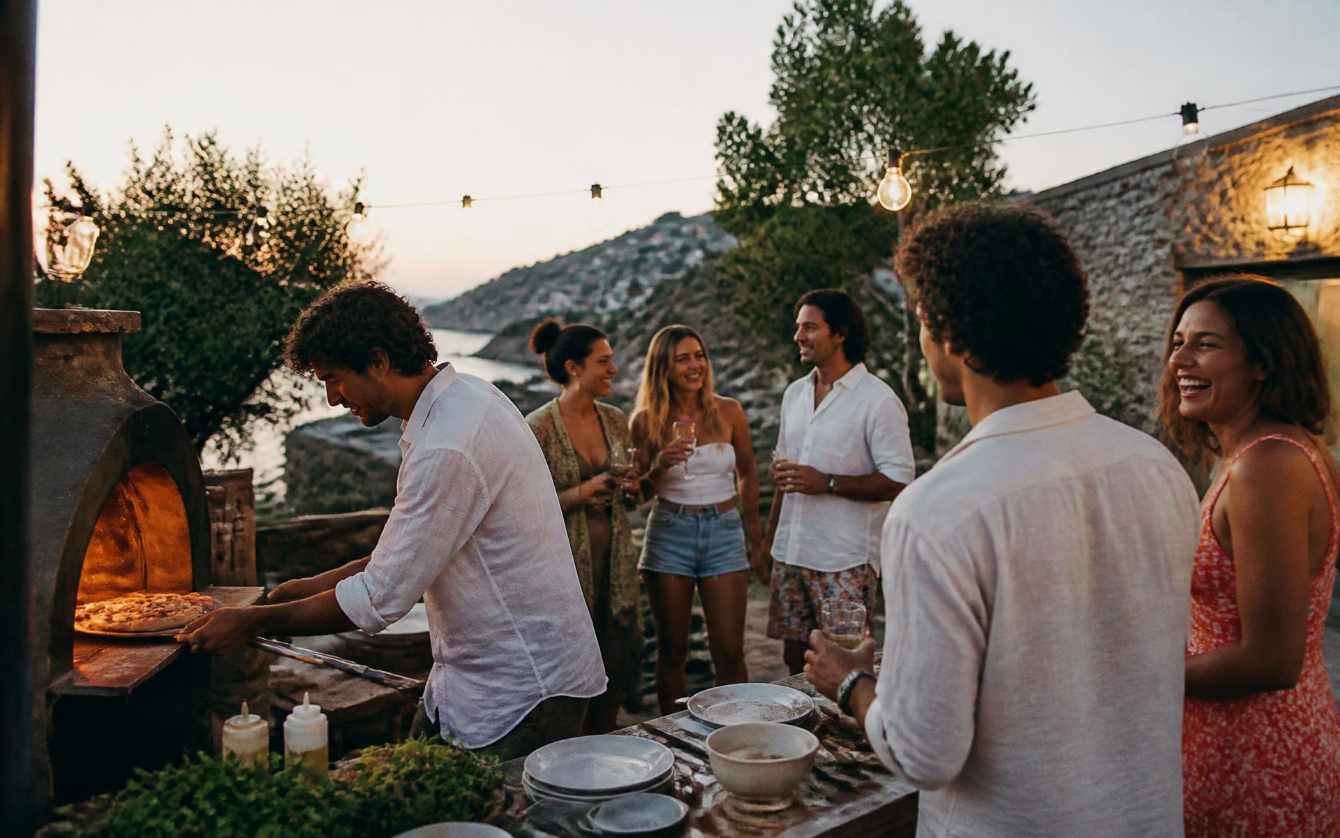 Amigos riendo alrededor de un horno de pizza en la terraza de una villa al atardecer