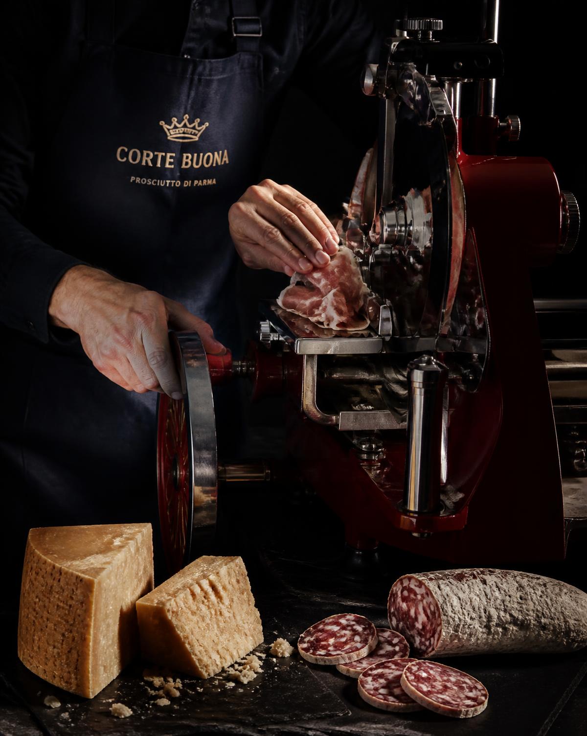 Corte de prosciutto en cortadora manual junto a quesos italianos